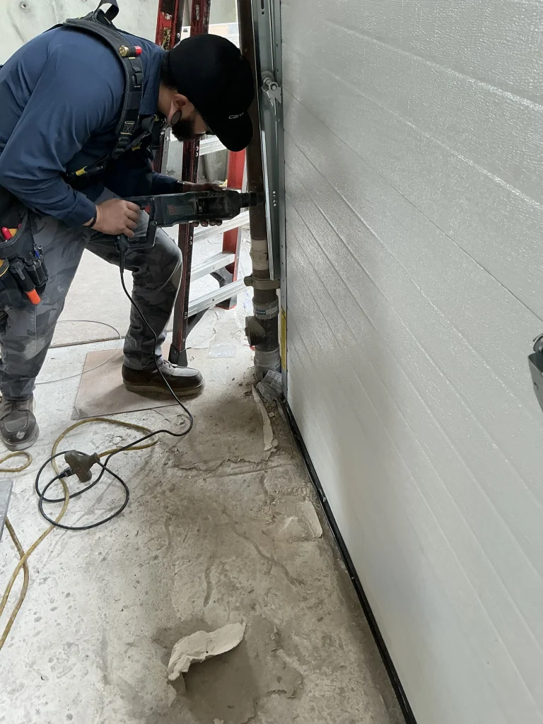 A technician using a heavy-duty rotary hammer drill to mount a steel track system to a concrete floor, illustrating the structural work that influences the average cost of a garage door.