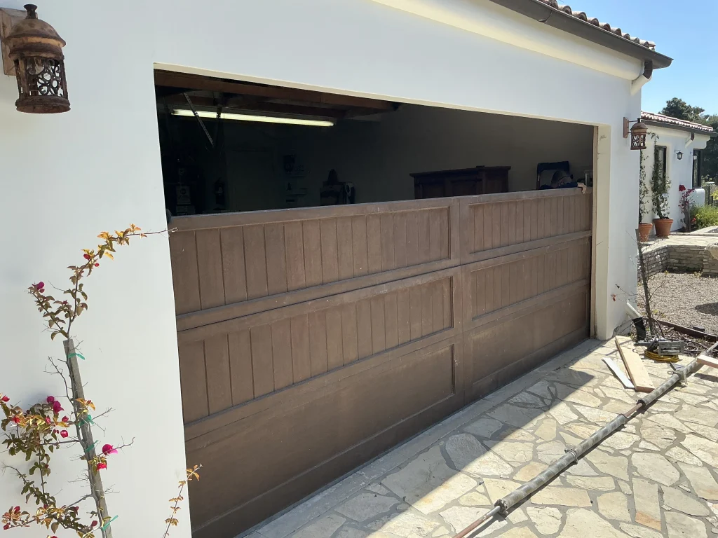 A brown wooden garage door stuck halfway open with components on the ground, showcasing a residential garage door repair.