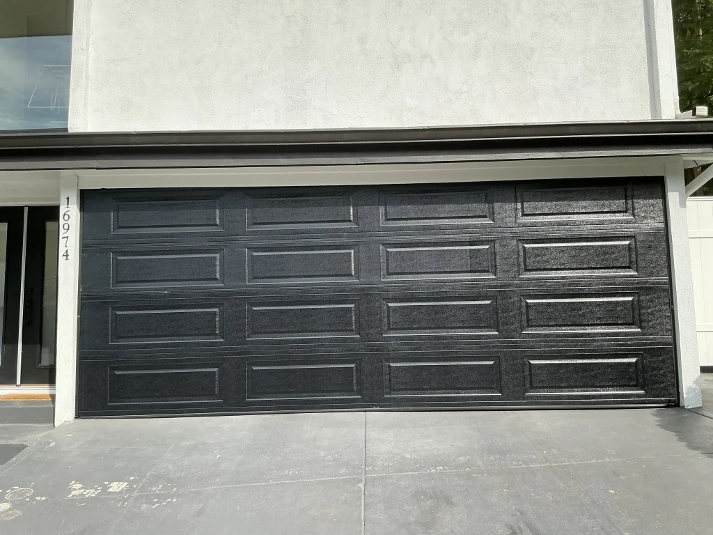 A wide, black double garage door with raised panels installed on a modern stucco home, showcasing standard garage door width.