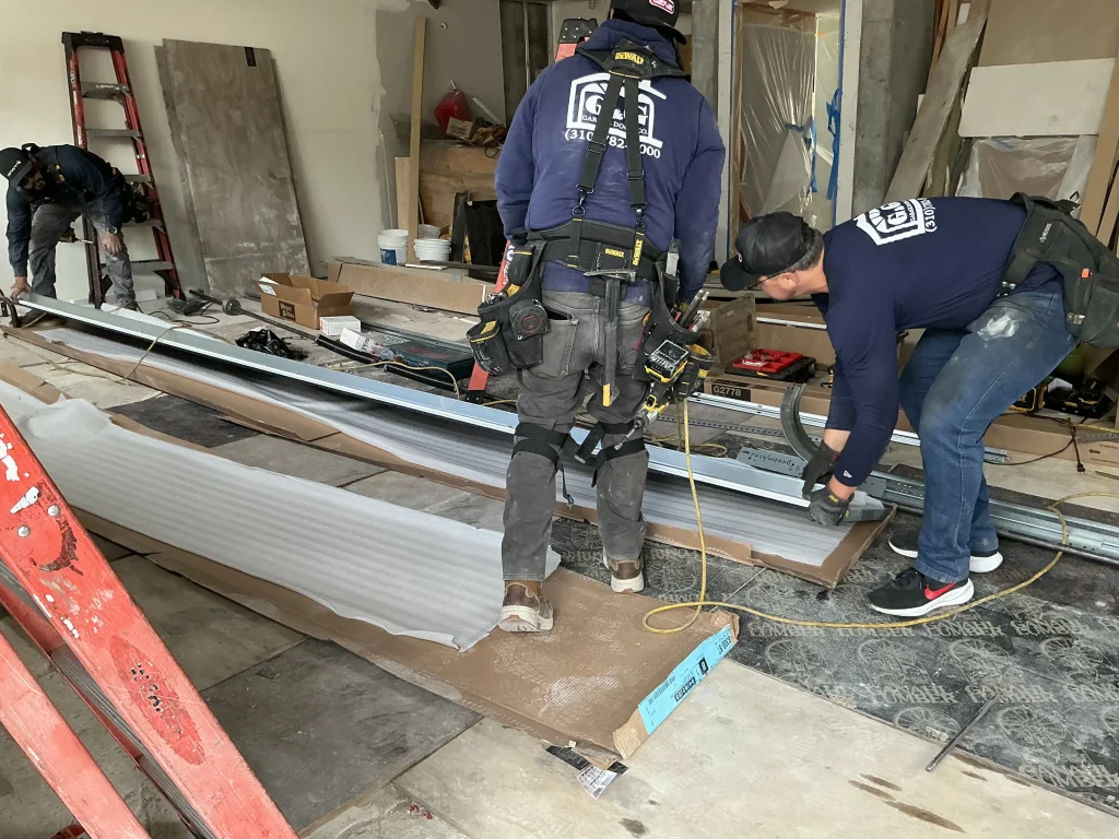 Three technicians assembling door panels and track components on a construction site floor, providing professional garage door services in Torrance, CA.