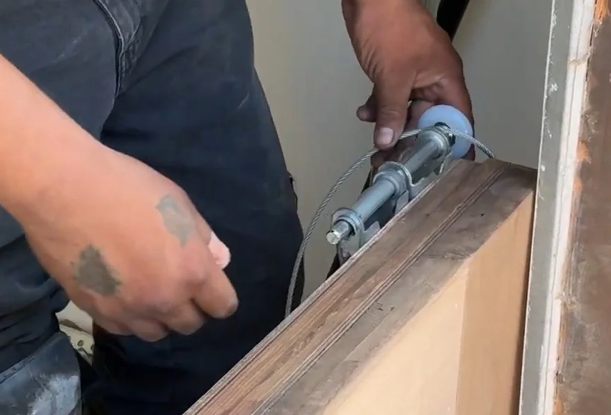 A close-up of a technician's hands threading a steel lift cable around a new white nylon roller fixture on a wood panel, demonstrating the installation of the best garage door rollers.