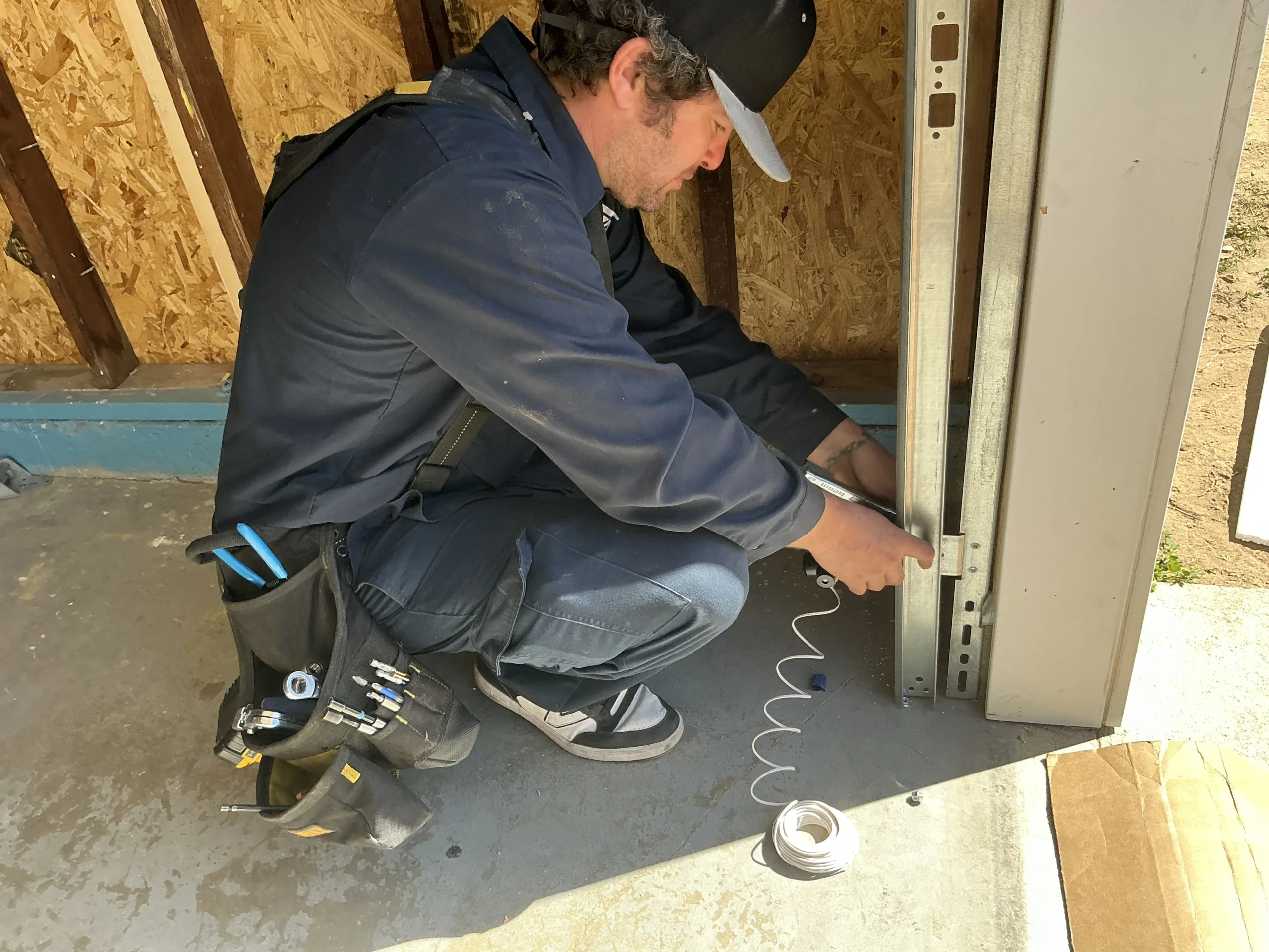 A technician crouching by a metal track to wire a safety photo-eye, demonstrating a professional garage door sensor repair.