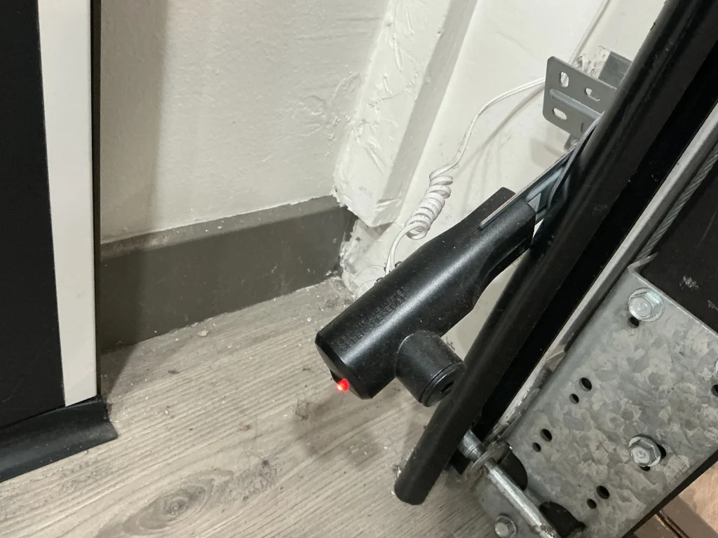 A close-up of a black safety photo-eye with a lit red LED mounted near the floor track, illustrating the installation of garage door sensors.