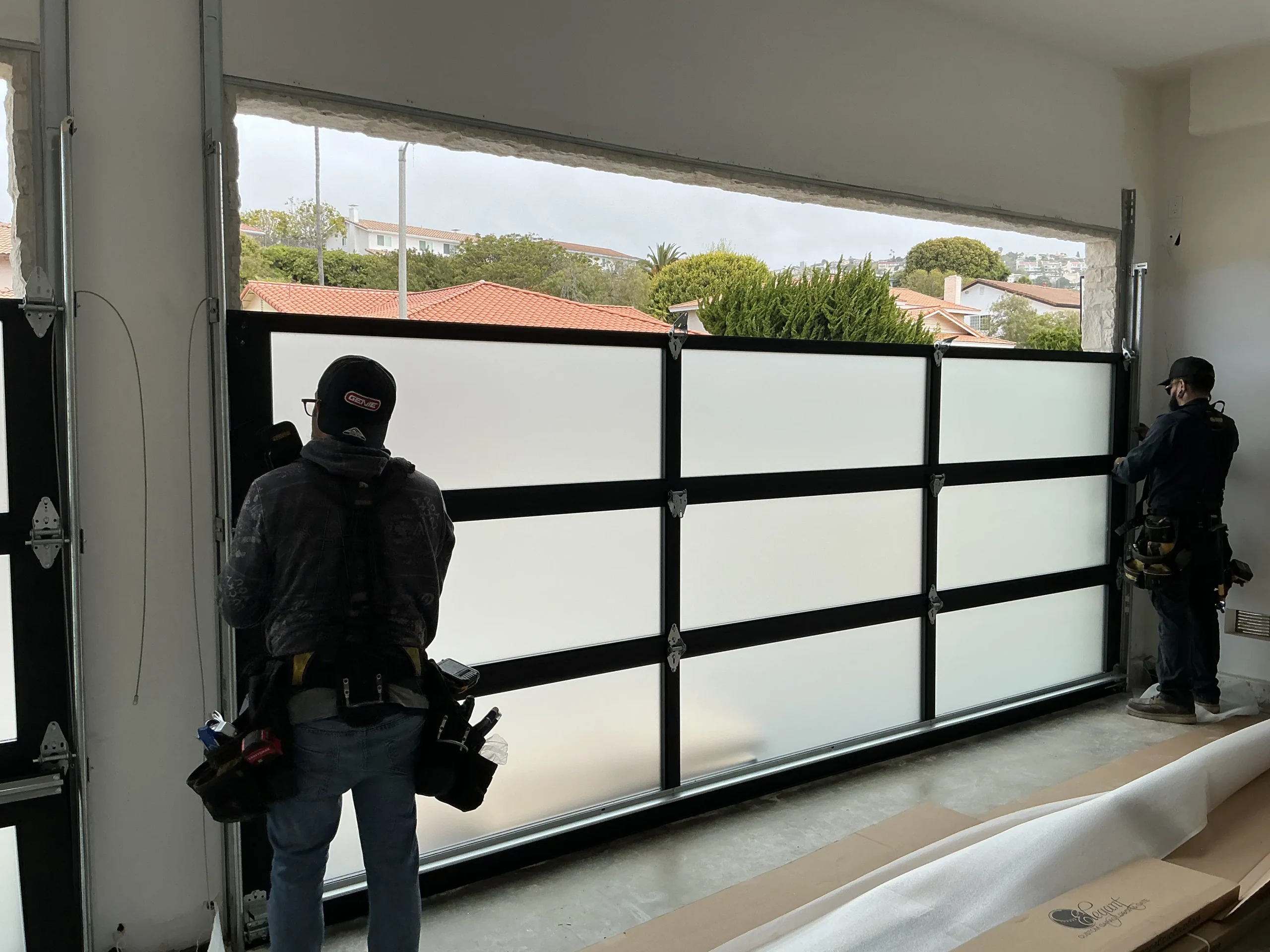 Two professional technicians adjusting the tracks of a modern black-framed frosted glass sectional door, demonstrating professional garage door maintenance near me.