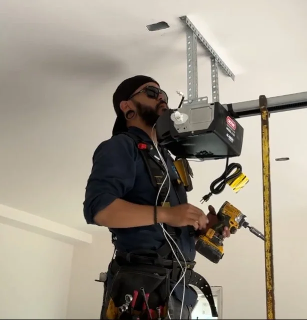 A technician holding a power drill while working on an overhead motor unit, performing a garage door opener battery replacement.