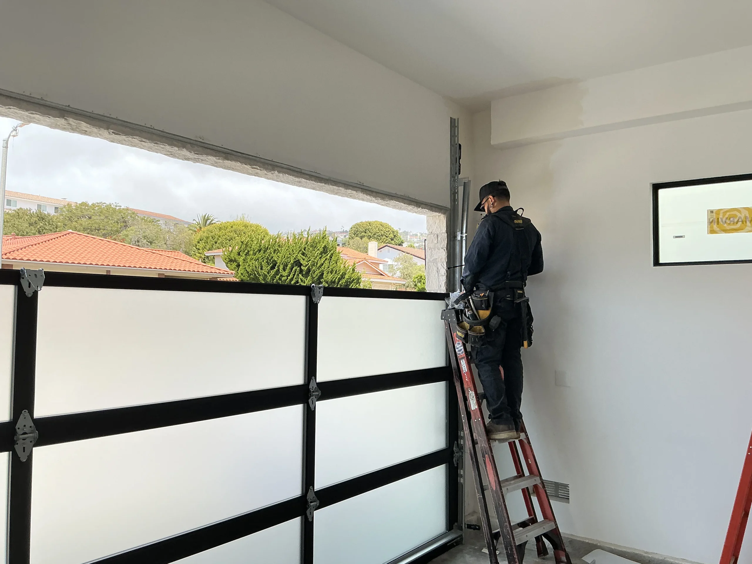 A professional technician standing on a ladder to fix a modern glass-panel track assembly, demonstrating the skilled repairs required for a damaged garage door.