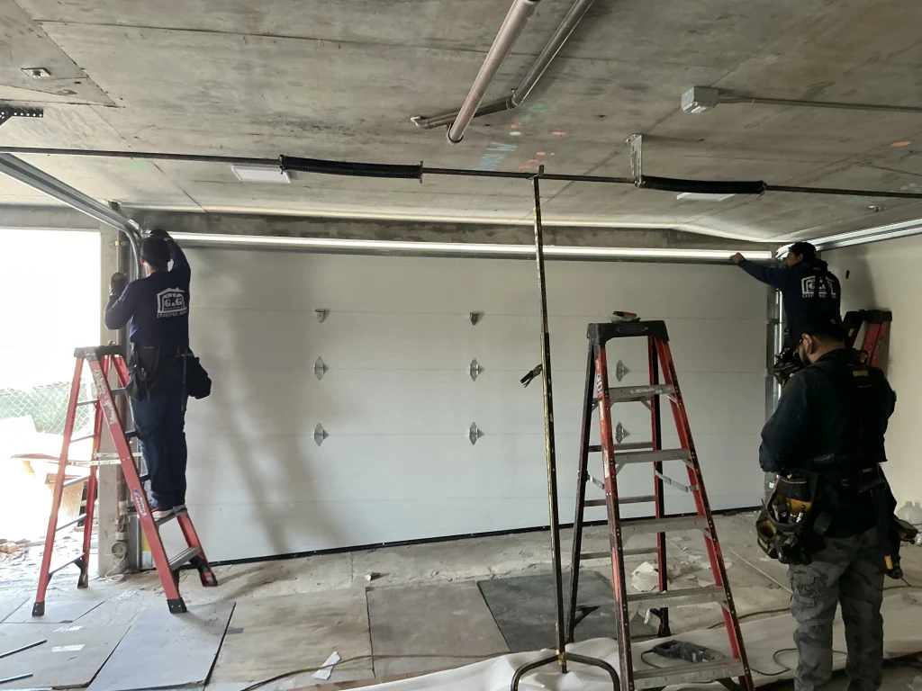 Three technicians working on ladders inside a concrete-walled garage to align a large white panel door system, highlighting a professional garage door repair service.