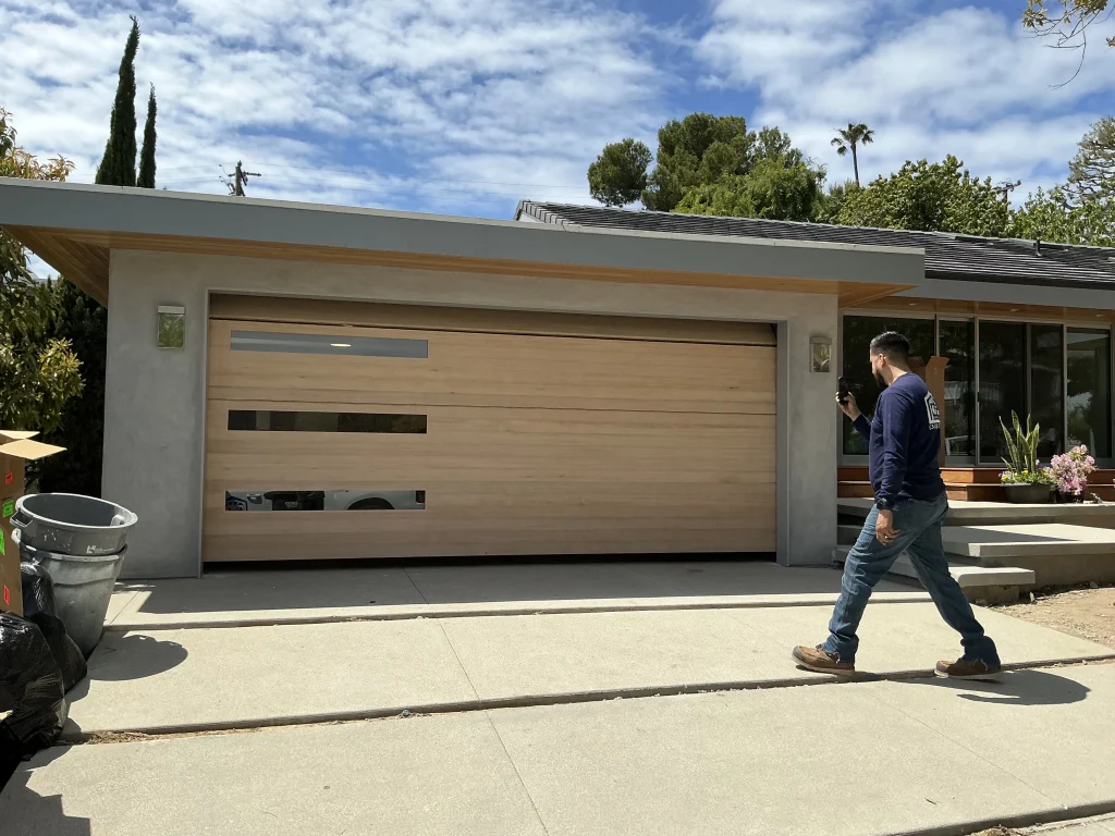 A service technician walking up a driveway toward a modern wood-paneled garage door with horizontal glass windows, ready to complete garage door repairs.