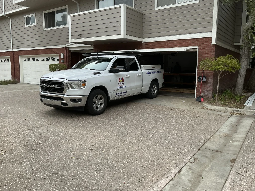 A white service truck parked in a residential driveway outside an open garage bay where a technician is performing preventative garage door maintenance.