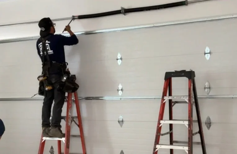 A professional technician standing on a ladder to carefully adjust an overhead torsion spring assembly, providing expert garage door services in Los Angeles, CA.