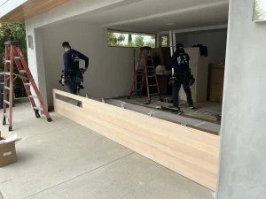 Two professional installers setting up lightweight wood-look panels on the driveway, demonstrating the layout of garage door sections.