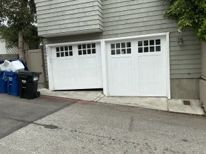 Alt Text: Two white carriage-style garage doors on a sloped driveway, with one door crooked, showing when you need to know how to open your garage door manually from outside.